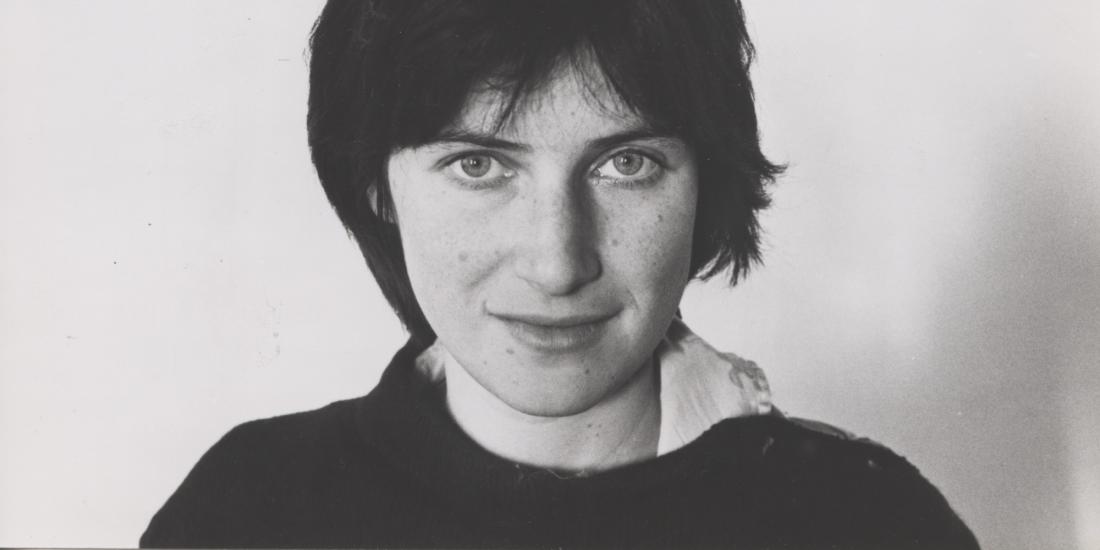 A black and white headshot of Chantal Akerman, who has one hand in her neck and stares straight into the camera, half-smiling.