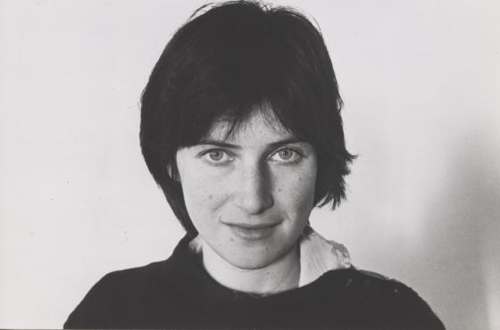 A black and white headshot of Chantal Akerman, who has one hand in her neck and stares straight into the camera, half-smiling.